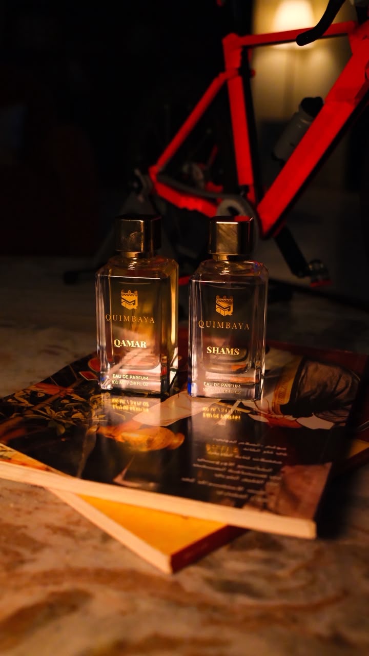 Two clear glass perfume bottles with gold caps and gold labels reading QAMAR & SHAMS, placed on top of open books in a dim indoor setting with a red bicycle frame blurred in the background.