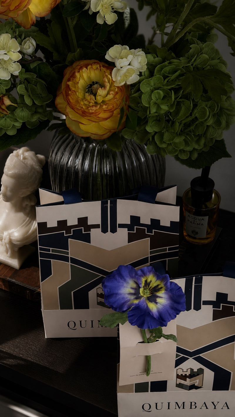 Quimbaya branded gift packaging bags with a its signature geometric navy, beige, and white pattern artwork inspired by Qasr Al Hosn, styled on a dark tabletop with a blue flower, a black ribbed vase of flowers, and a small bottle in the background.
