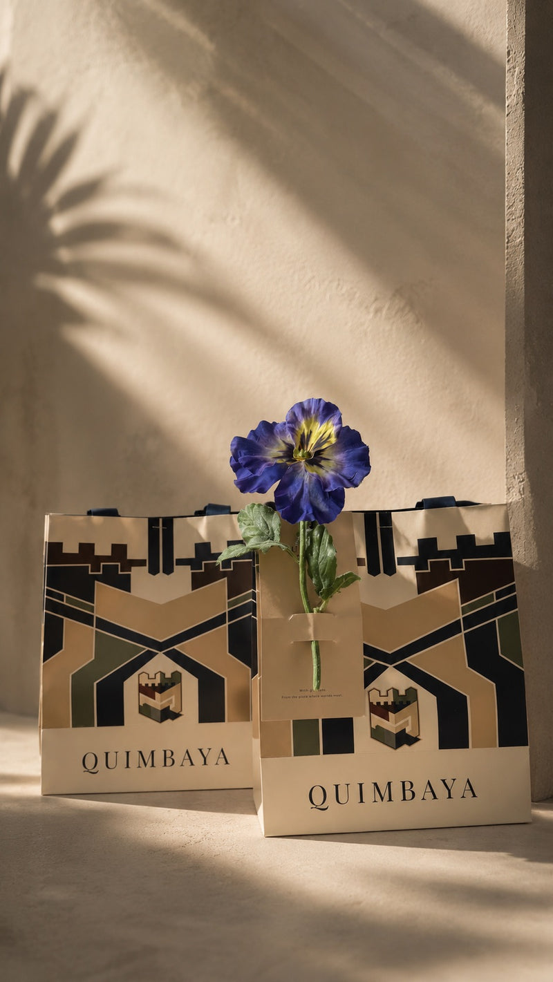 Two Quimbaya branded shopping bags with signature geometric bags on a warm beige surface with dramatic shadow casting.